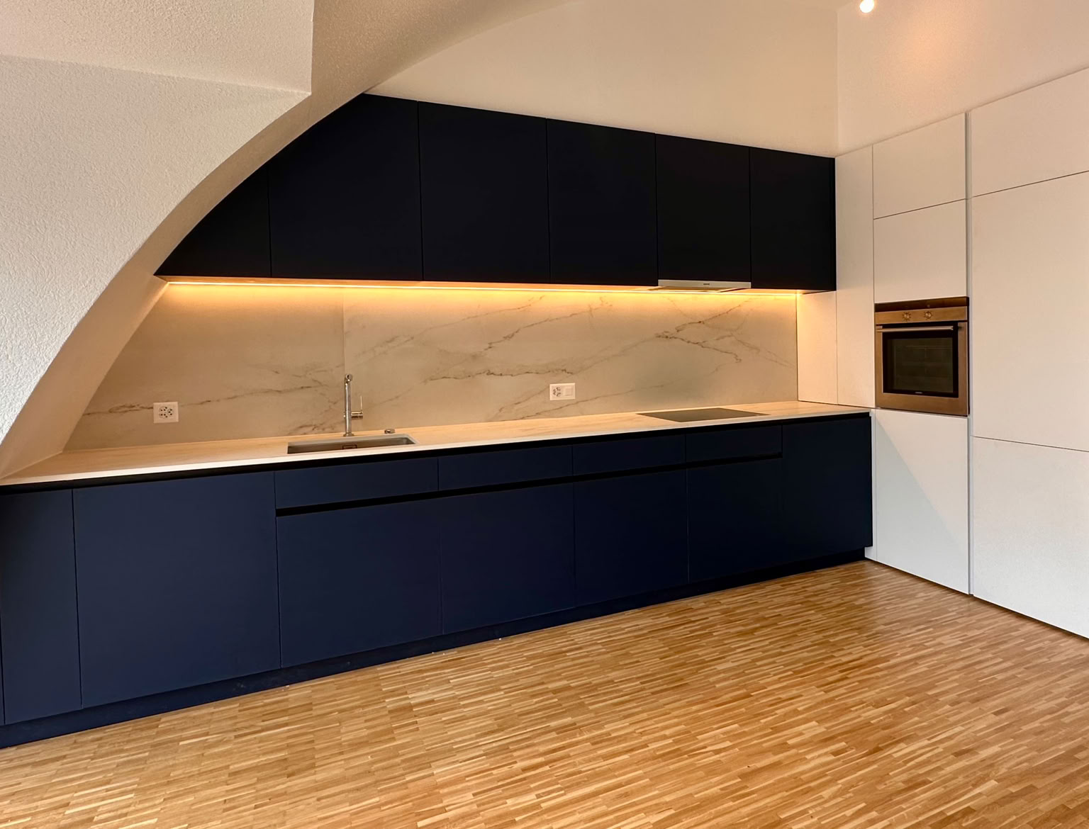 Cuisine moderne avec des armoires bleue nuit, un plan de travail clair, un éclairage sous les armoires, un évier, et un four encastré contre un mur blanc arrondi,Cuisine moderne avec des armoires bleue nuit, un plan de travail clair, un éclairage LED  encastré contre un mur blanc arrondi, concept Biolley Pollini