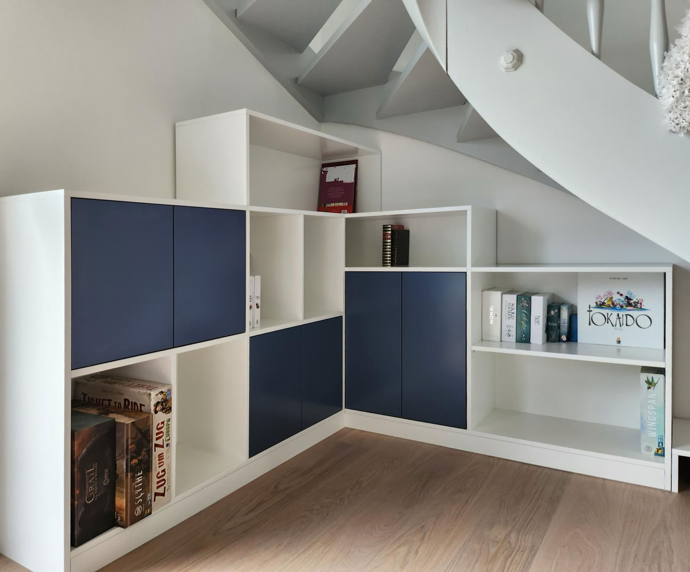 Étagère moderne en angle avec des compartiments ouverts et fermés, contenant des livres et des objets décoratifs, située sous un escalier.